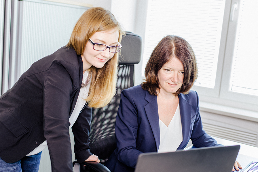 Zwei Frauen in Businesskleidung schauen gemeinsam auf einen Laptop.