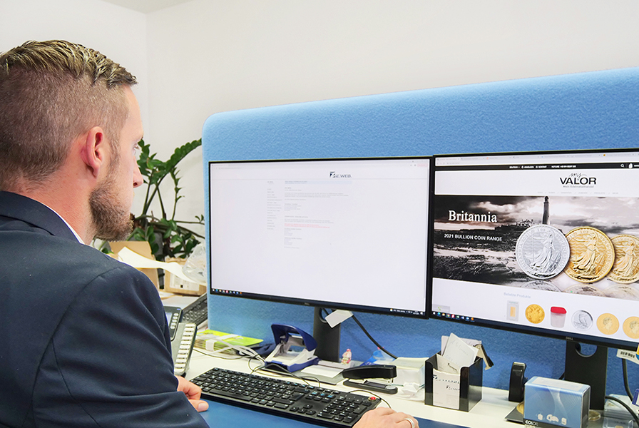 Ein Mitarbeiter blickt konzentriert auf zwei Bildschirme, auf denen Informationen über Münzen und Edelmetalle zu sehen sind. 